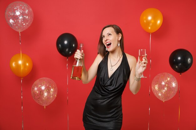 Joyeuse jeune femme en petite robe noire célébrant, tenant un verre, une bouteille de champagne sur des ballons à air de fond rouge vif. Saint Valentin, concept de fête de vacances de maquette d'anniversaire de bonne année.