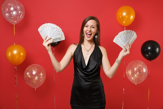 Joyeuse jeune femme en petite robe noire célébrant la tenue d'un paquet de dollars, de l'argent liquide sur des montgolfières à fond rouge vif. Concept de fête de vacances de maquette d'anniversaire de bonne année de la femme.