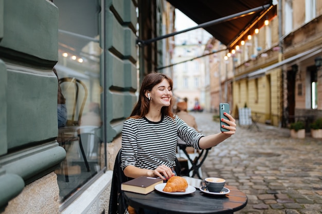 Joyeuse jeune femme prenant un selfie sur un smartphone moderne assis à la terrasse d'un café confortable