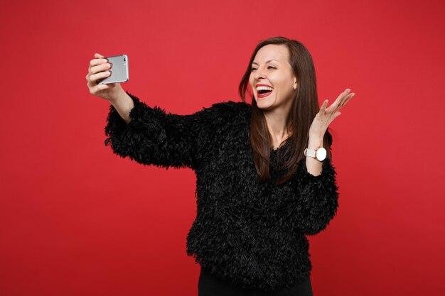 Joyeuse jeune femme en pull de fourrure noire écartant les mains, faisant une photo de selfie sur un téléphone portable isolé sur fond de mur rouge vif. Les gens émotions sincères, concept de style de vie. Maquette de l'espace de copie.