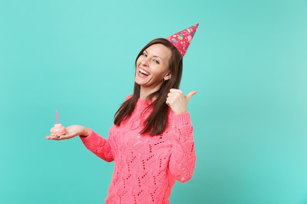 Joyeuse jeune femme en pull rose tricoté, chapeau d'anniversaire pointant le pouce de côté tenir dans le gâteau à la main avec une bougie isolée sur fond bleu portrait en studio. Concept de mode de vie des gens. Maquette de l'espace de copie.