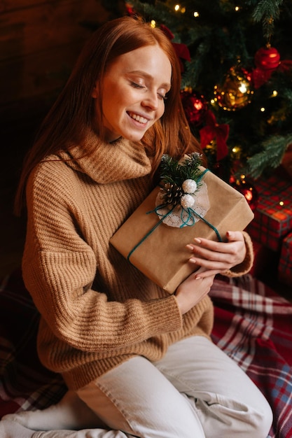 Joyeuse jeune femme rousse aux yeux fermés tenant une boîte-cadeau à la main reçue pour Noël