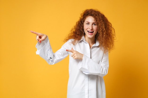 Joyeuse jeune femme rousse en chemise blanche posant isolée sur un mur orange jaune