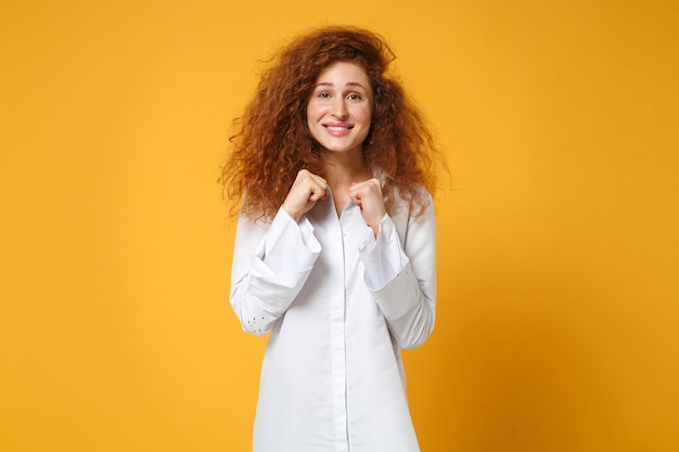 Joyeuse jeune femme rousse souriante en chemise blanche décontractée posant isolée sur un mur orange jaune