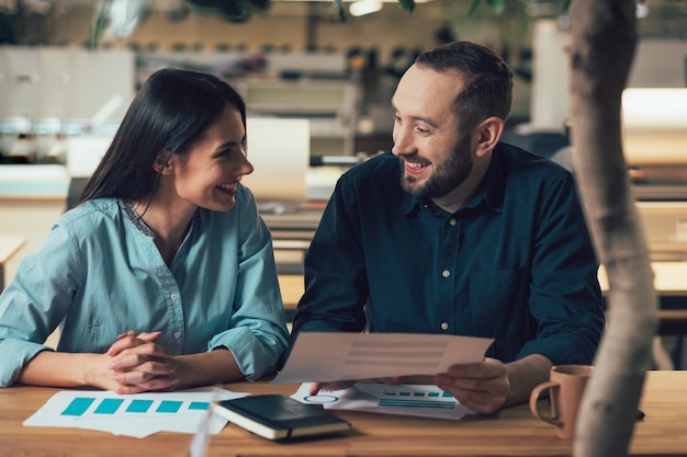 Joyeuse jeune femme souriante à un homme joyeux assis à table avec lui et discutant des chartes graphiques devant eux