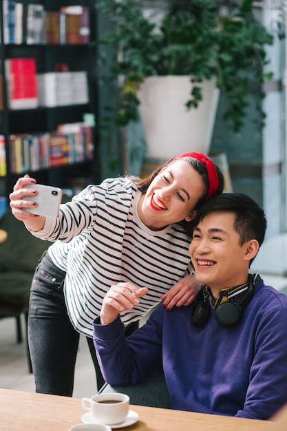 Joyeuse jeune femme souriante et se penchant vers un homme positif à ses côtés tout en tenant un smartphone et en prenant un selfie