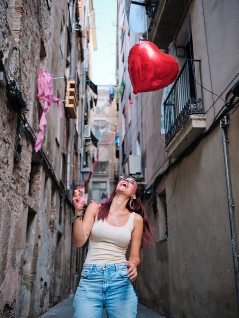 Joyeuse jeune femme tenant un ballon coeur rouge tout en se tenant à l'extérieur dans la rue.