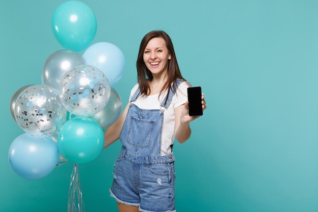 Joyeuse jeune femme tenant un téléphone portable avec un écran vide vierge, célébrant avec des ballons à air colorés isolés sur fond de mur bleu turquoise. Fête d'anniversaire, concept d'émotions de personnes.