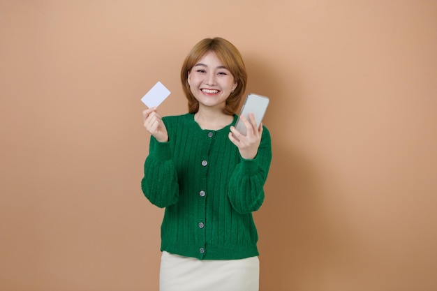 Joyeuse jeune femme utilisant un téléphone portable tient une carte bancaire de crédit isolée sur fond beige