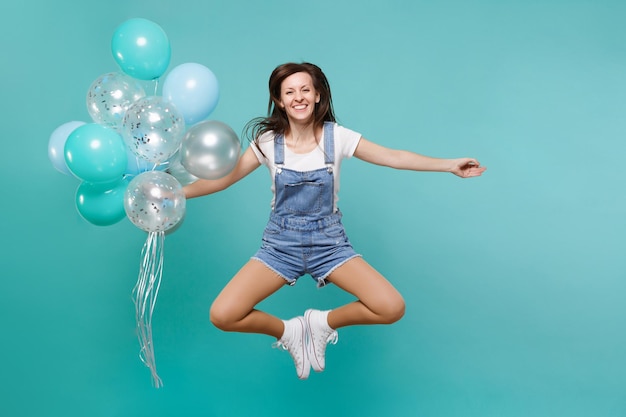 Joyeuse jeune femme en vêtements en denim écartant les mains, sautant haut, célébrant et tenant des ballons à air colorés isolés sur fond bleu turquoise. Fête d'anniversaire, concept d'émotions de personnes.