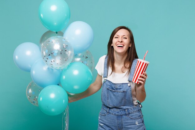 Joyeuse jeune femme en vêtements en denim tenant une tasse en plastique de cola ou de soda célébrant avec des ballons à air colorés isolés sur fond bleu turquoise. Fête d'anniversaire, concept d'émotions de personnes.