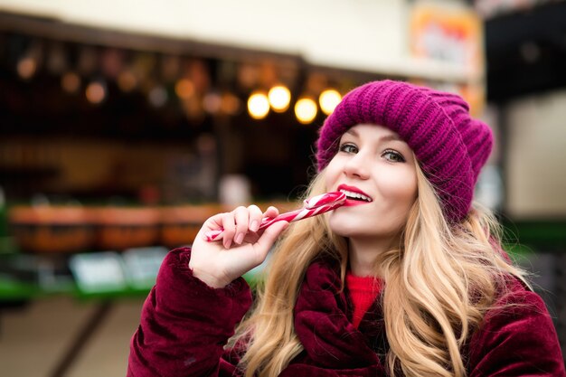 Joyeuse jeune fille blonde au bonnet rouge posant avec une canne en bonbon à la foire de Noël