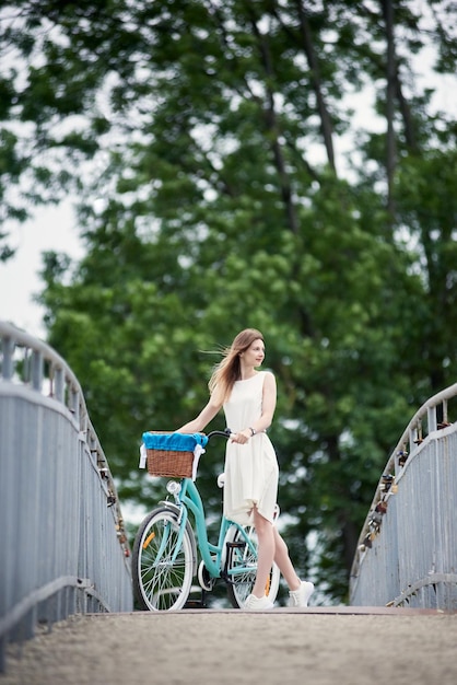 Joyeuse jeune fille debout avec un vélo bleu vintage et profitant de la nature sur le pont