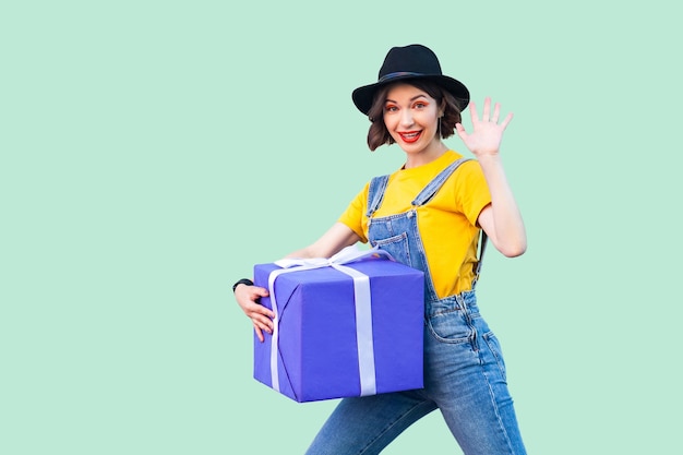 Joyeuse jeune fille heureuse en vêtements hipster en salopette en jean et chapeau noir debout et essayant de tenir une grosse boîte-cadeau lourde et géante et vous saluant, en montrant cinq. Studio shot, fond vert, isolé