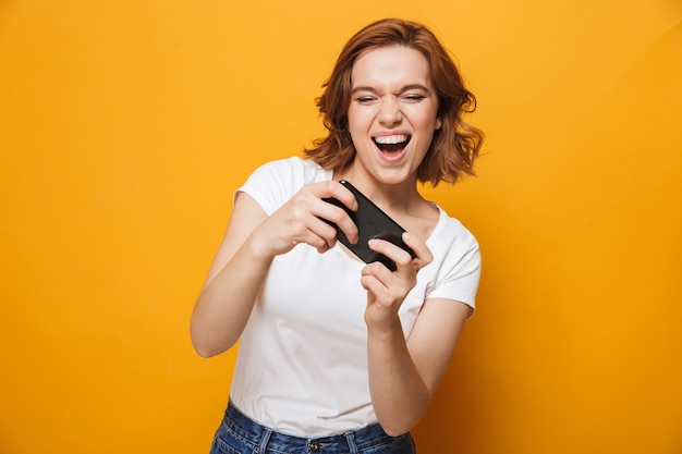 Joyeuse jeune fille portant un t-shirt debout isolée sur un mur jaune, jouant à des jeux sur téléphone portable