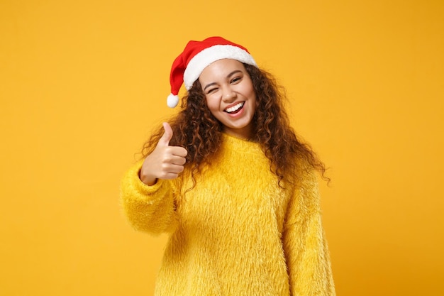 Joyeuse jeune fille de Santa afro-américaine en pull de fourrure, chapeau de Noël isolé sur fond jaune. Bonne année 2020 concept de vacances de célébration. Maquette de l'espace de copie. Montrant le pouce vers le haut, clignotant.