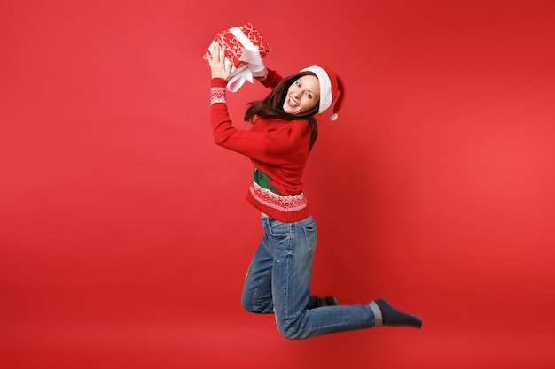 Joyeuse jeune fille de Santa en chapeau de Noël sautant tenant une boîte rouge avec un cadeau, présente isolée sur fond de mur rouge vif. Bonne année 2019 concept de fête de vacances célébration. Maquette de l'espace de copie.