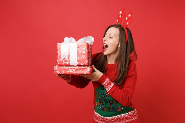 Joyeuse jeune fille de Santa dans des cornes de cerf décoratives amusantes tenant des boîtes rouges avec des cadeaux, gardant la bouche grande ouverte isolée sur fond rouge. Bonne année 2019 concept de fête de vacances célébration.