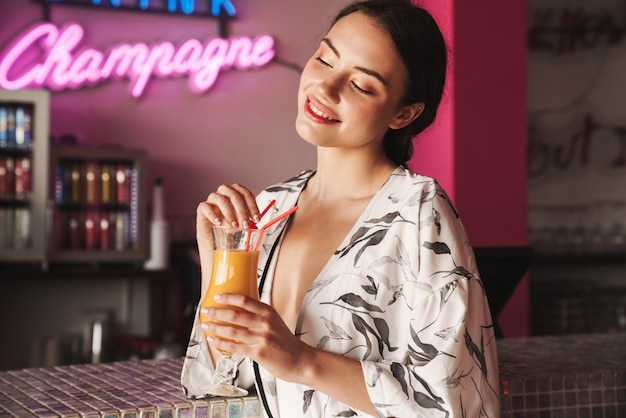 joyeuse jeune jolie femme à la fête sur la plage avec un mur coloré à led lumineux dans un café-bar buvant un cocktail.