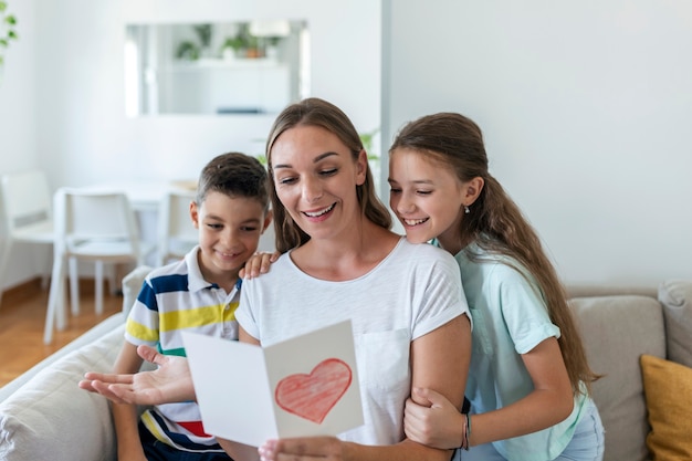 Joyeuse jeune mère et petits enfants assis sur un canapé et s'embrassant en lisant les souhaits et félicitations pour les vacances en carte postale présentée à la maison