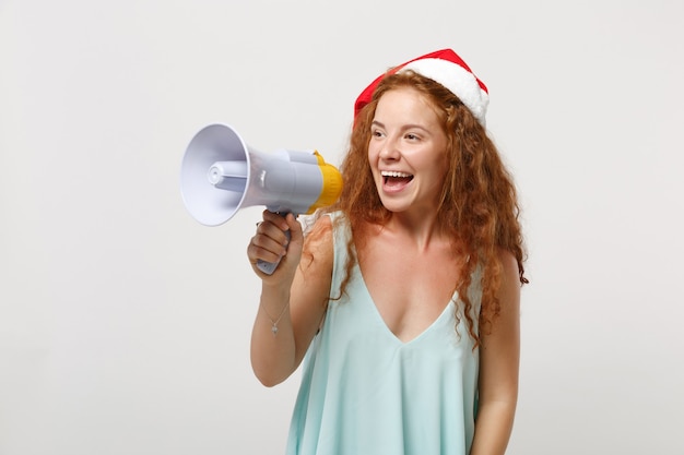 Joyeuse jeune rousse Santa girl dans des vêtements légers, chapeau de Noël isolé sur fond de mur blanc en studio. Concept de vacances de célébration de bonne année 2020. Maquette de l'espace de copie. Crier dans le mégaphone.