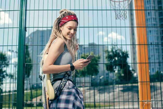 Joyeuse jolie dame debout à côté de la clôture à mailles de chaîne et souriant tout en regardant l'écran de son appareil moderne