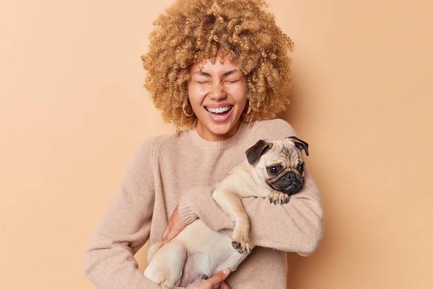 Joyeuse jolie femme aux cheveux bouclés, propriétaire d'un animal de compagnie, porte un chien carlin qui s'amuse avec son animal domestique préféré qui rit et a de bonnes relations vêtu d'un pull décontracté isolé sur fond de studio beige.