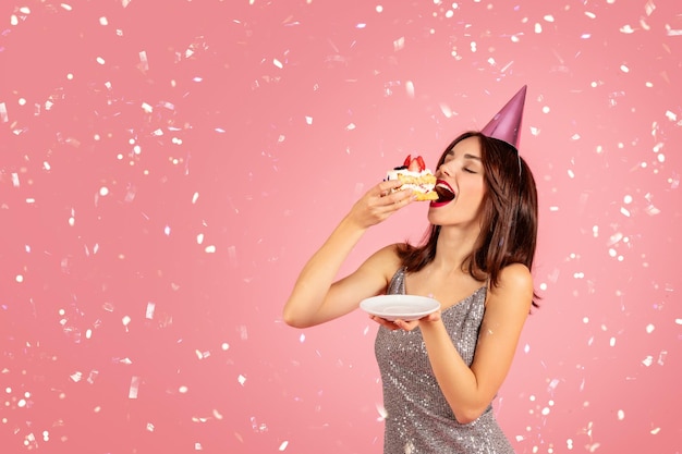 Joyeuse jolie femme brune caucasienne du millénaire dans une robe de chapeau de fête mange du gâteau