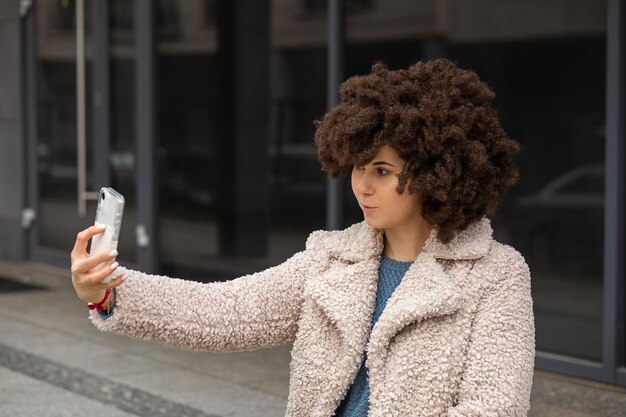 Joyeuse jolie femme mignonne. Portant une fausse perruque Afro Coiffure. Prend un selfie, sourit joyeusement. Faire du contenu photo pour le blog. Porter un manteau en fausse fourrure. Style de rue