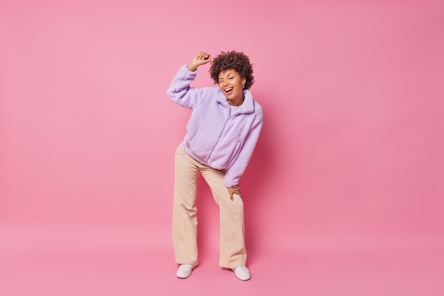 Joyeuse jolie femme millénaire a une humeur enjouée cligne des yeux l'œil lève le bras se sent très heureux porte un manteau de fourrure pantalon à fond de cloche et des baskets pose pour faire une photo isolée sur le mur rose du studio