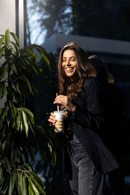 Joyeuse jolie femme se tient près d'un pot de fleurs tient une boisson au café dans ses mains ferma les yeux et sourit rêveusement