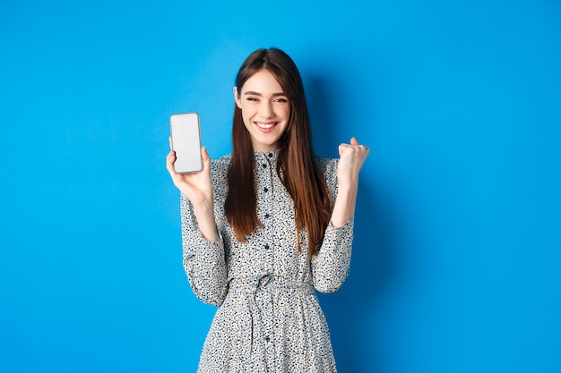 Joyeuse jolie fille chantant, gagnant sur téléphone mobile, montrant l'écran du smartphone vide et la pompe à poing, souriant et célébrant, bleu.