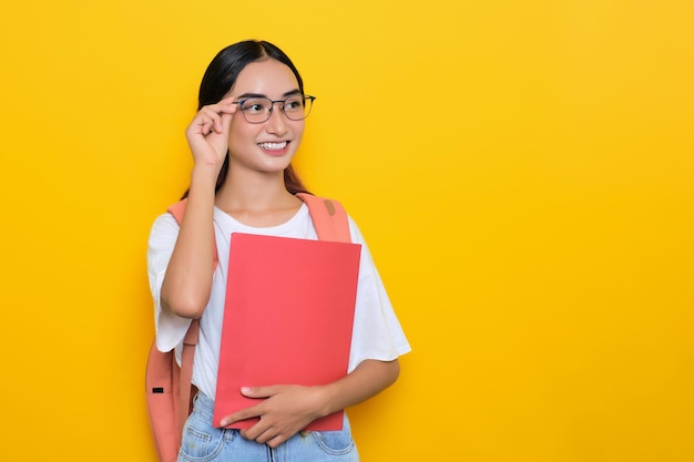 Joyeuse jolie jeune étudiante portant un sac à dos et des lunettes tenant des livres isolés sur fond jaune