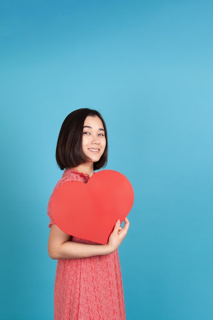 Joyeuse, joyeuse jeune femme asiatique en robe rouge tenant grand coeur de papier rouge comme fan