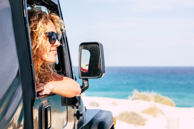 Joyeuse liberté heureuse nrautiful jeune femme caucasienne regarde à l'extérieur de la voiture à la plage avec ciel bleu et scène de l'océan