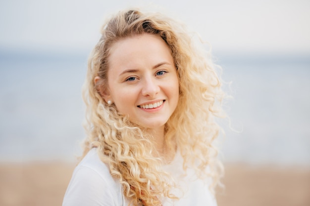 Joyeuse, optimiste, jeune femme a le sourire brillant, les cheveux bouclés, respire l’air pur au bord de la mer.