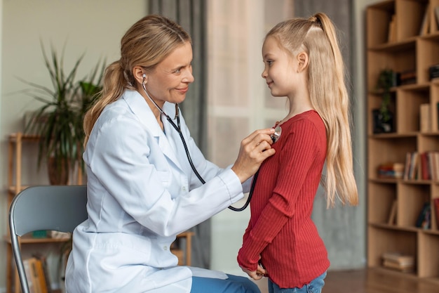 Joyeuse pédiatre à l'écoute du rythme cardiaque de la fille avec stéthoscope lors de l'examen à la maison vue latérale