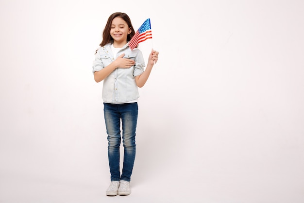 Joyeuse petite fille animée tenant le drapeau américain et touchant sa poitrine tout en exprimant son bonheur