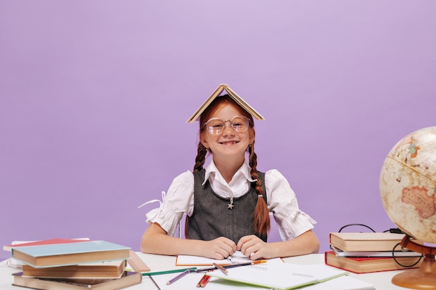 Joyeuse petite fille aux cheveux rouges en uniforme scolaire élégant souriant et assis au bureau avec des livres et des cahiers sur fond lilas