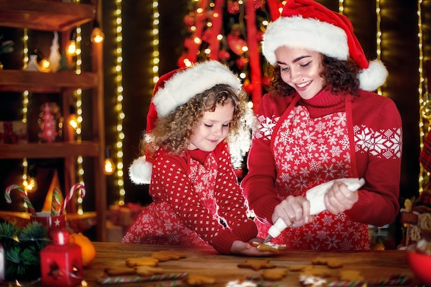 Joyeuse petite fille bouclée mignonne et sa sœur aînée en chapeaux de santa cuisiner des biscuits de Noël