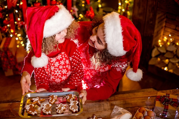 Joyeuse petite fille bouclée mignonne et sa sœur aînée en chapeaux de santa cuisiner des biscuits de Noël