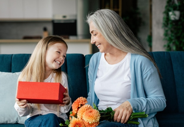 Joyeuse petite fille caucasienne aime ouvrir une boîte-cadeau avec une vieille dame avec un bouquet de fleurs dans le salon