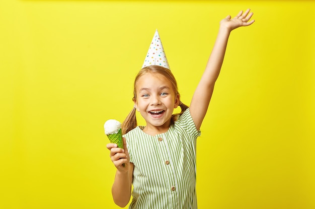 joyeuse petite fille dans un chapeau d'anniversaire a une humeur joyeuse en mangeant de la crème glacée se dresse sur jaune isolé