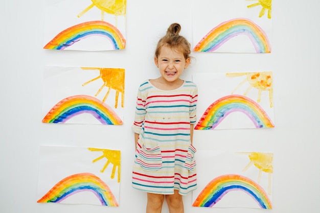 Joyeuse petite fille énergique dans une chemise rayée à côté de six peintures similaires de soleil et d'arc-en-ciel qu'elle a peintes Posant devant le mur