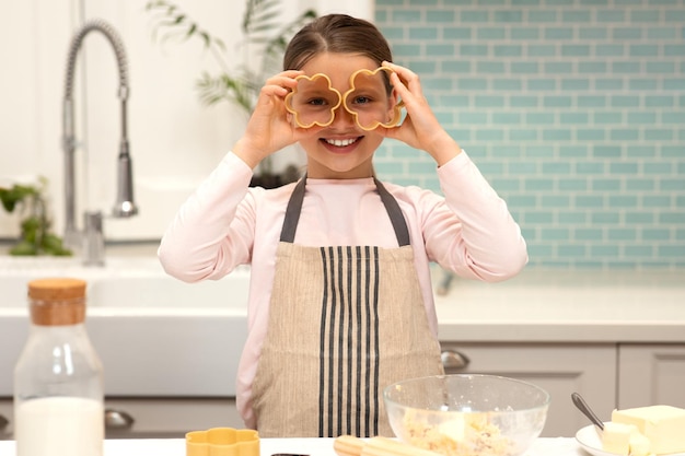 Joyeuse petite fille européenne en tablier fait des biscuits met des emporte-pièces aux yeux s'amuse à l'intérieur de la cuisine