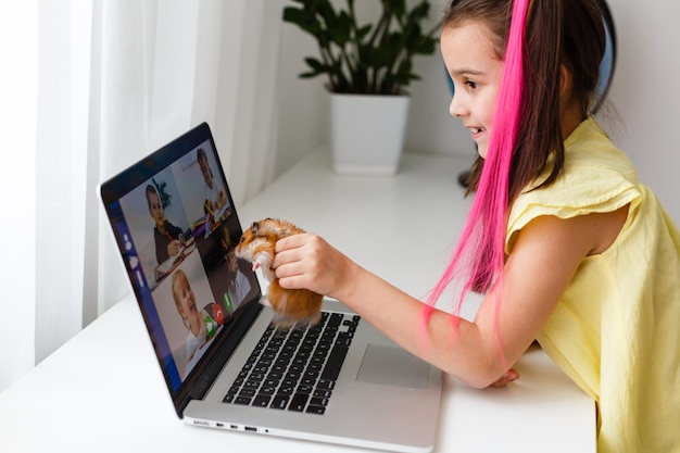 Joyeuse petite fille avec un hamster pour animaux de compagnie utilisant un ordinateur portable étudiant via un système d'apprentissage en ligne à la maison. Formation à distance ou à distance