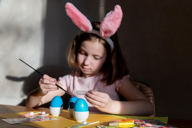 Joyeuse petite fille heureuse s'amusant et décorant des oeufs de Pâques pour les vacances de printemps assis à table dans une cuisine confortable