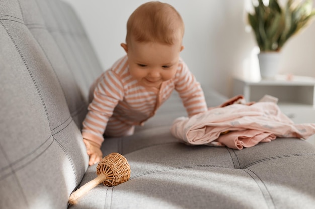 Joyeuse petite fille joue avec des jouets en bois et des vêtements à la maison. Enfant d'un an, rampant sur le canapé avec un jouet à la main, pose au soleil.