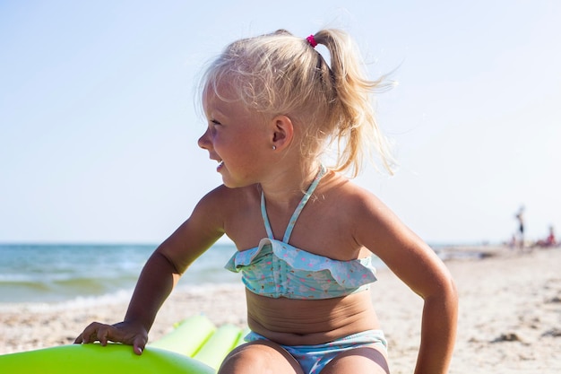 Joyeuse petite fille en maillot de bain est assise sur un matelas gonflable sur la plage.