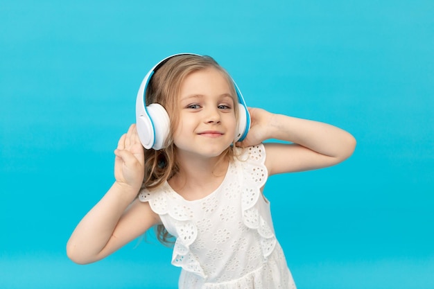 Joyeuse petite fille mignonne avec un casque écoutant de la musique dans une robe blanche en coton sur fond bleu dans le studio et un espace souriant pour le texte